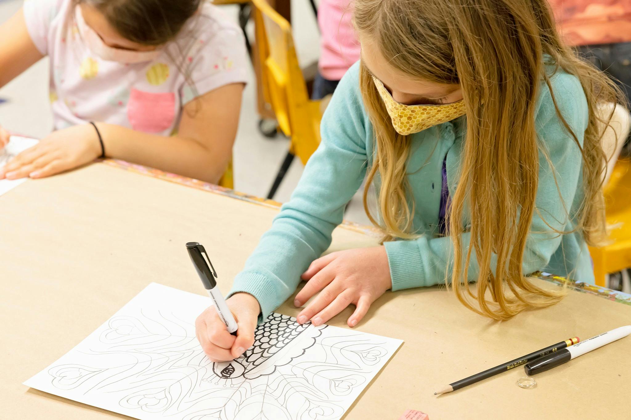 Child in a mask drawing on paper with a marker in a classroom