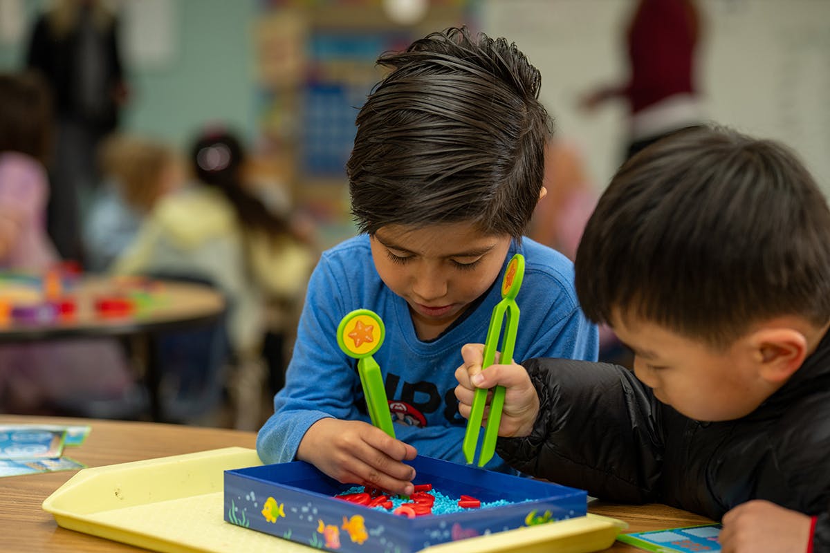 Two children playing with small objects and green tools in a classroom