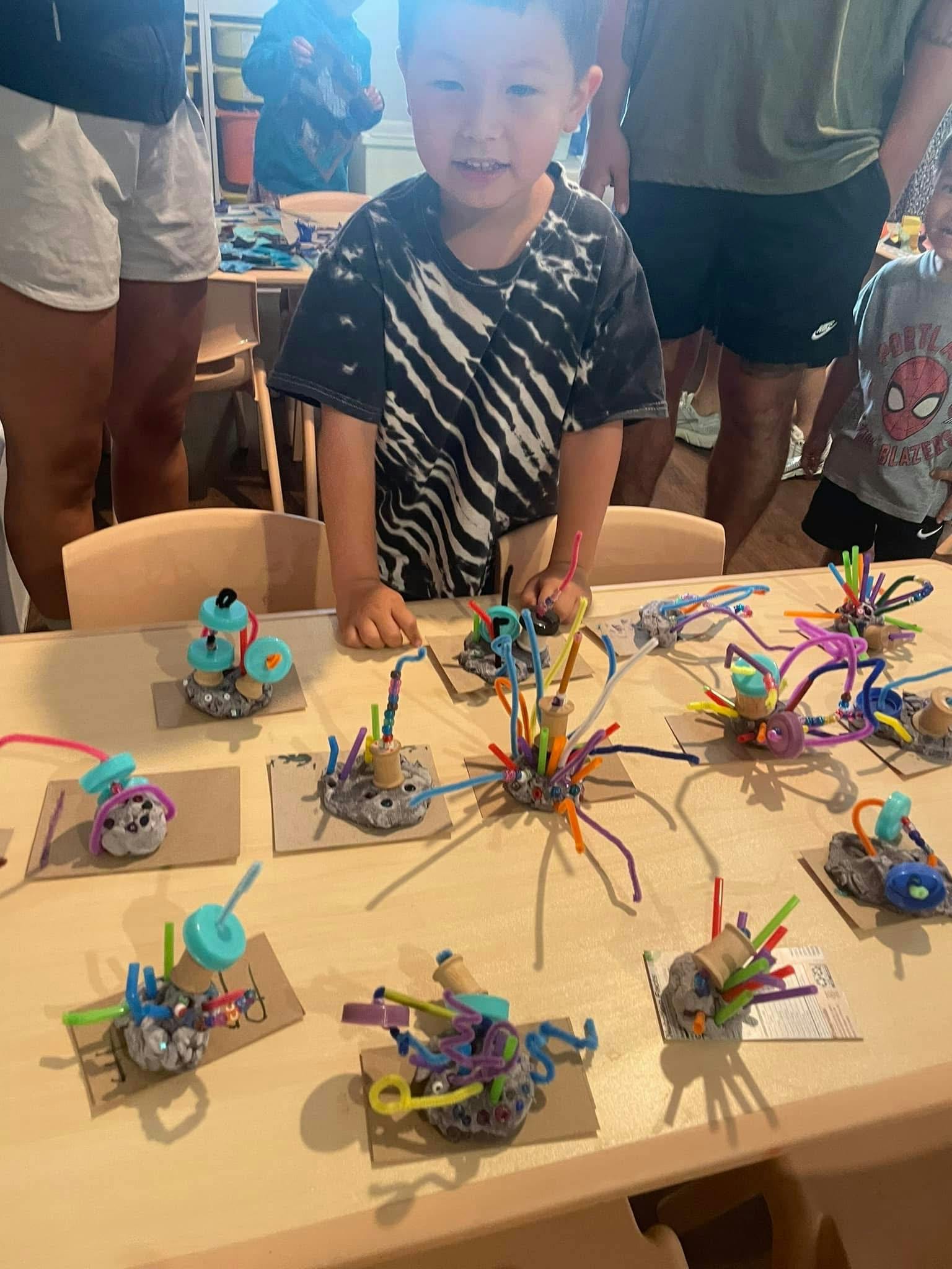 Child standing at a table displaying numerous colorful craft creations