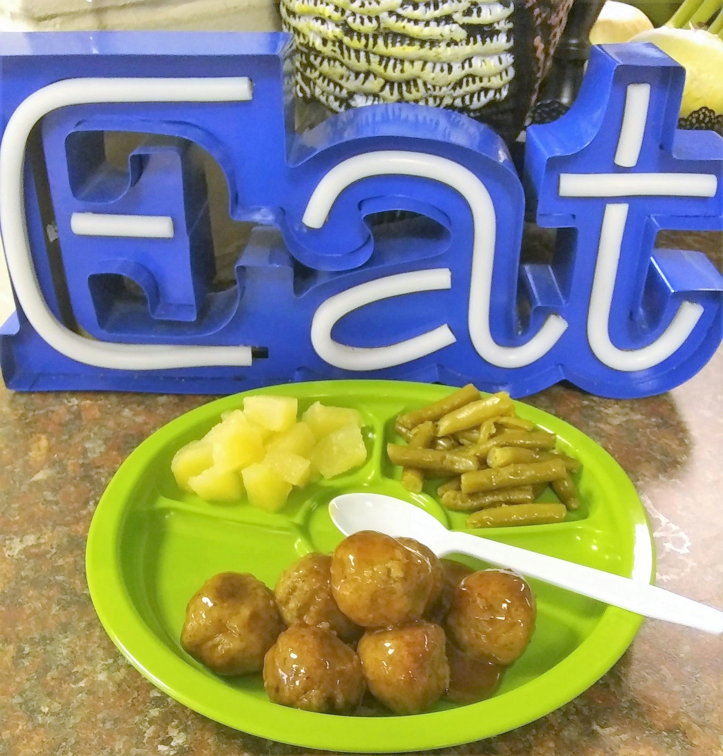 Plate of meatballs, green beans, and pineapple next to a blue "EAT" sign