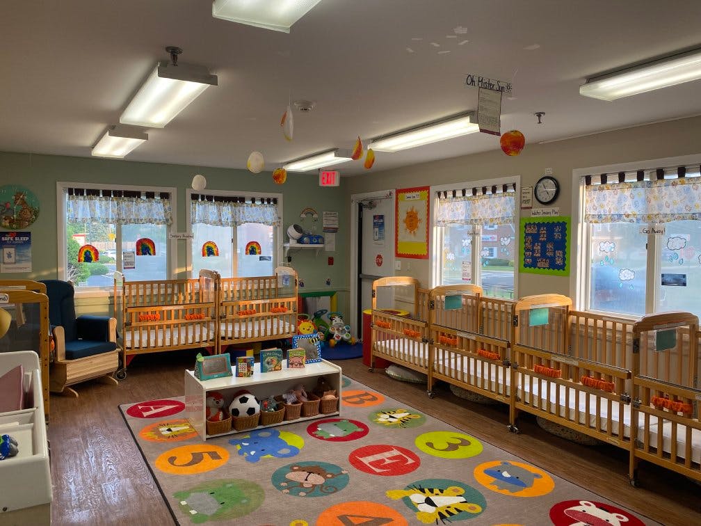 A brightly lit nursery room with multiple wooden cribs along the walls and a colorful rug