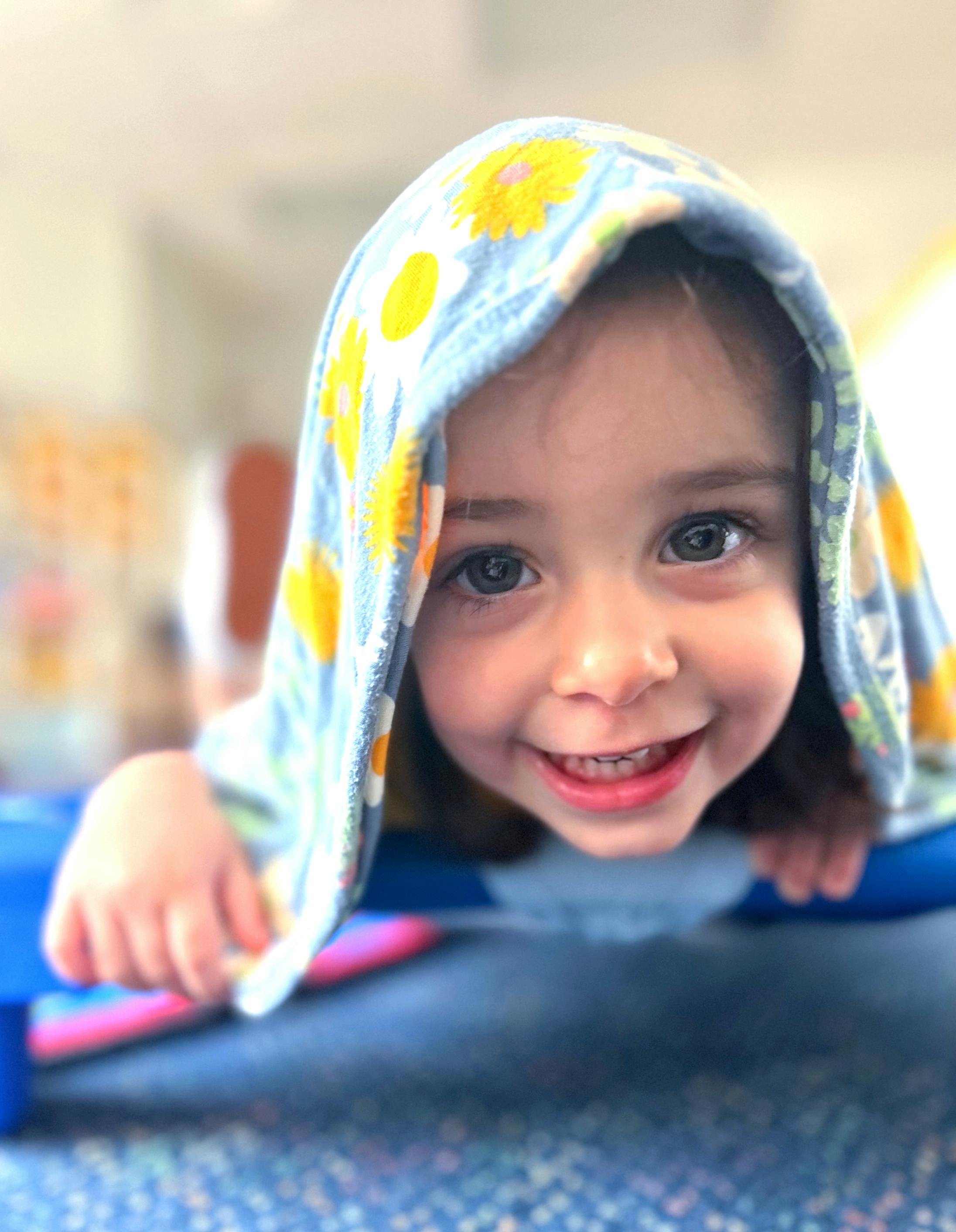 Child smiling with a blanket over their head, looking directly at the camera