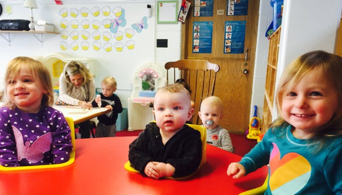 Three children sitting at a red table, one adult and child in background