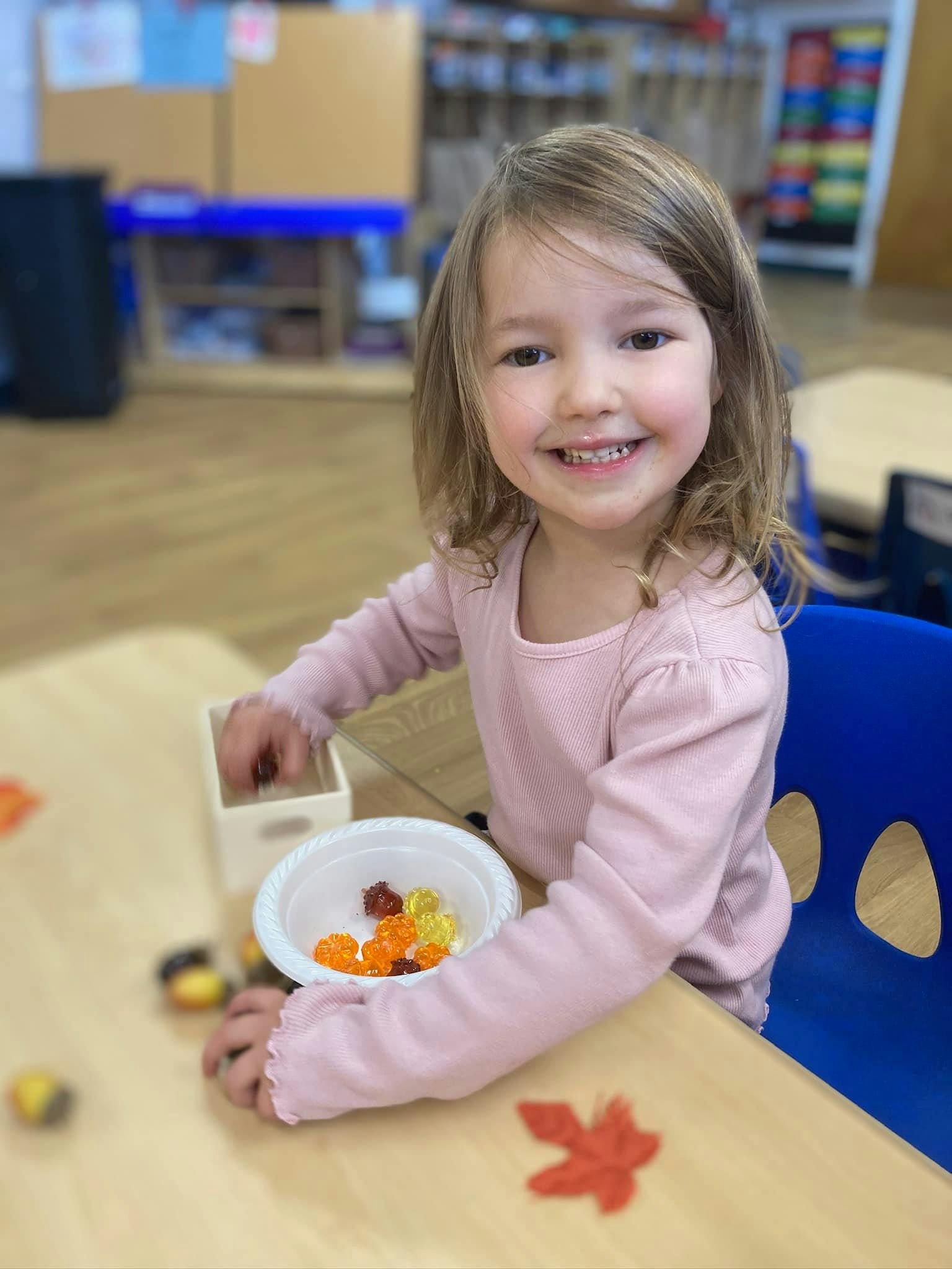 Child smiling while playing with small objects at a table in a classroom