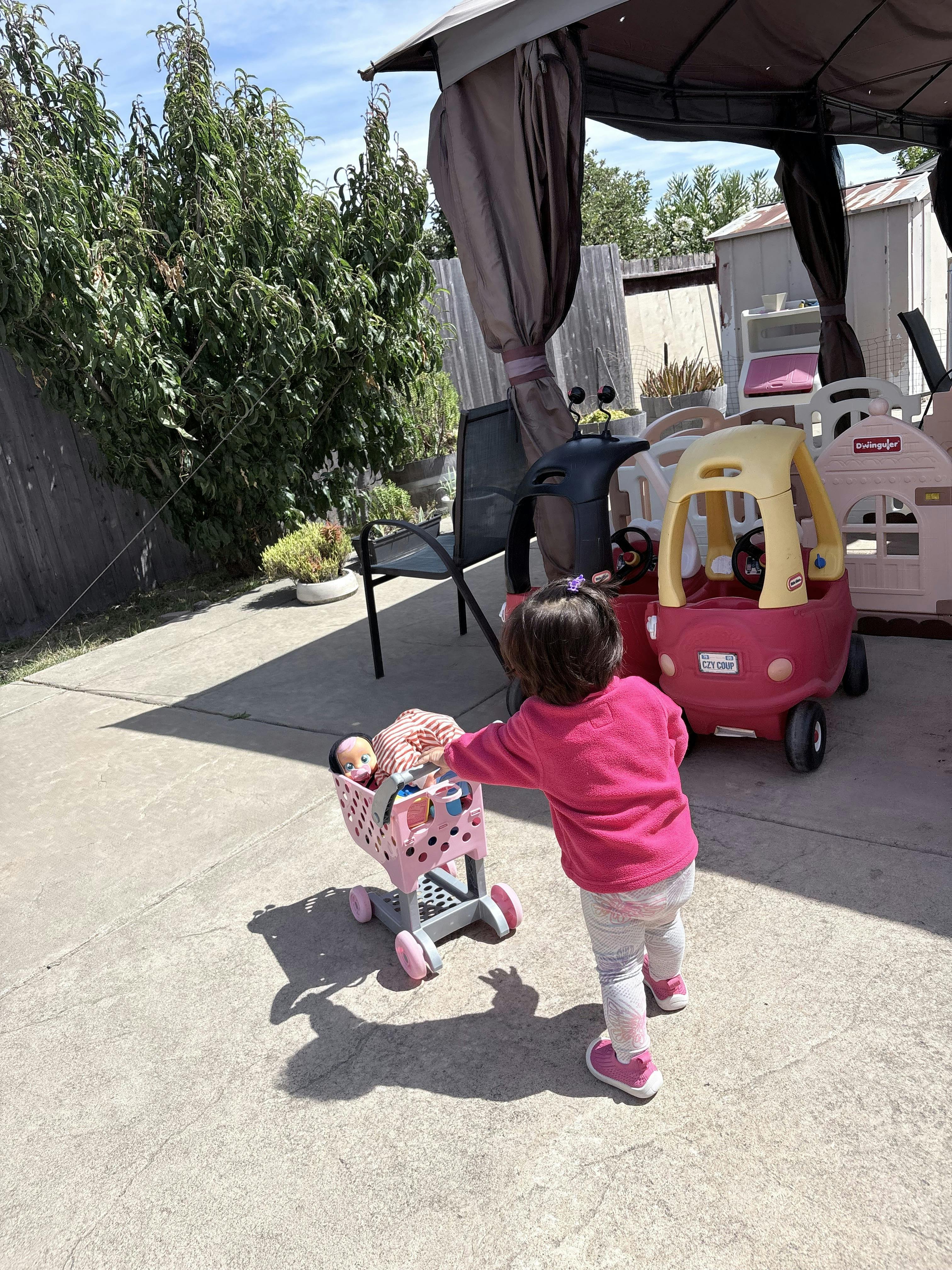 Child pushing a toy shopping cart with a doll in a patio area