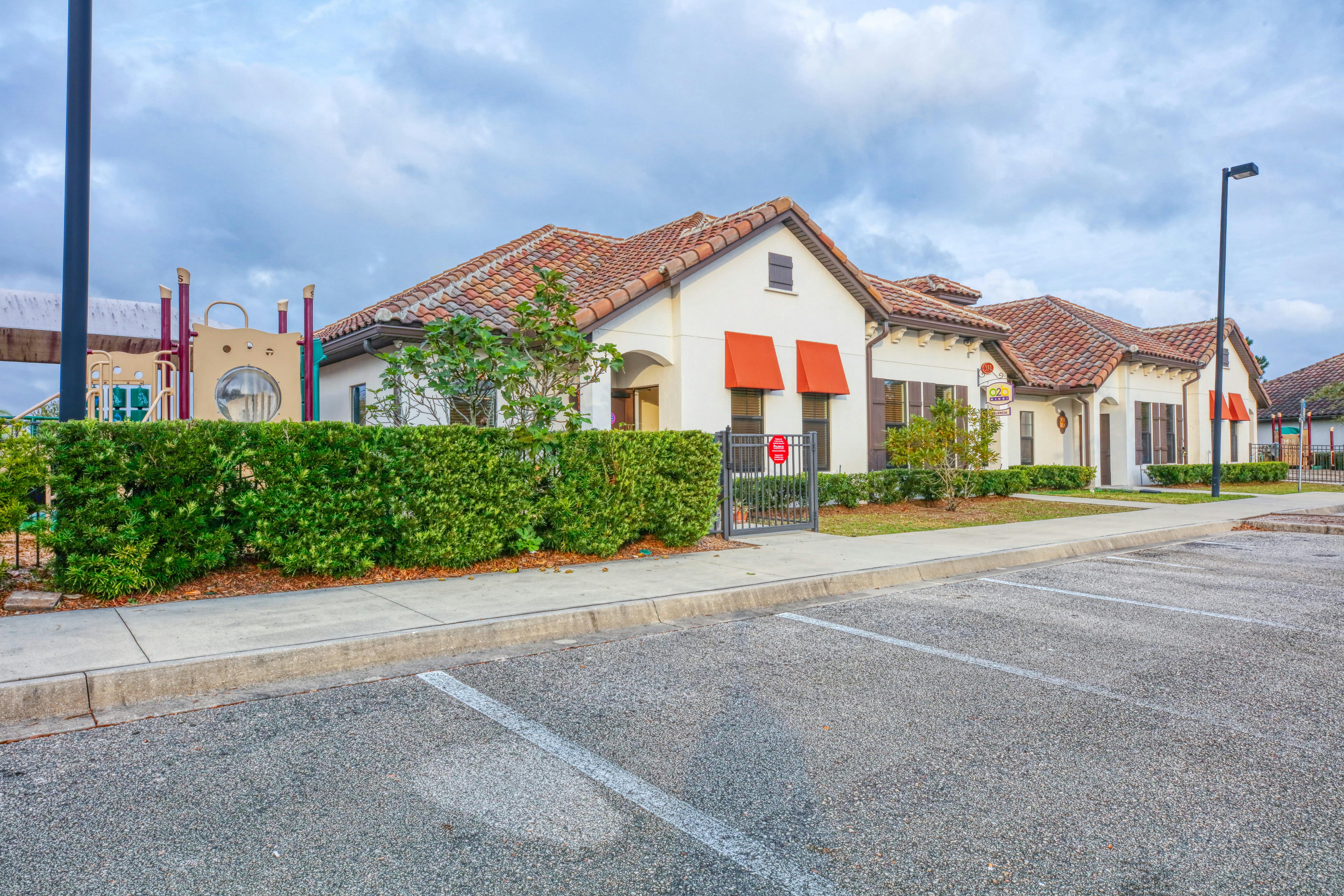 A beige building with red tile roofs and orange awnings next to a parking lot and playground