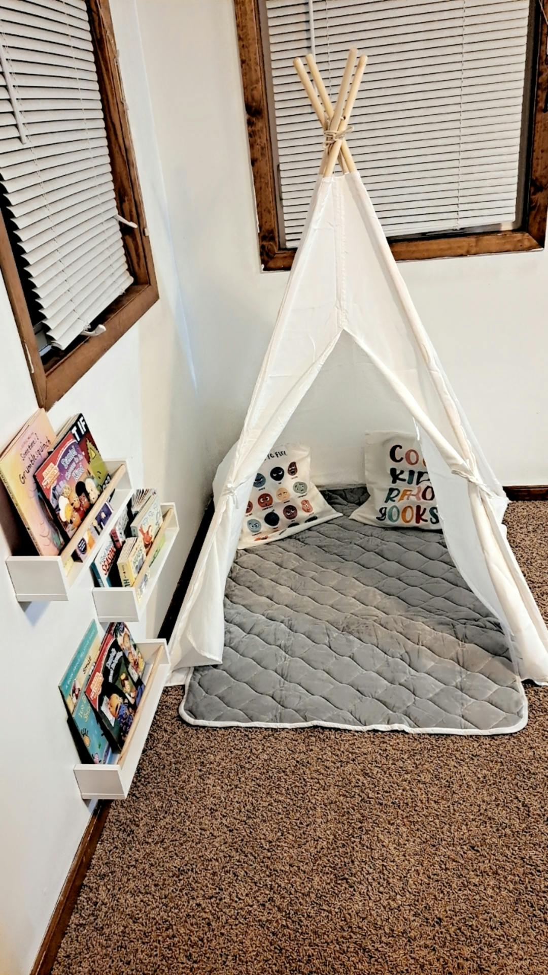 White teepee with a gray mat and pillows next to wall-mounted book shelves