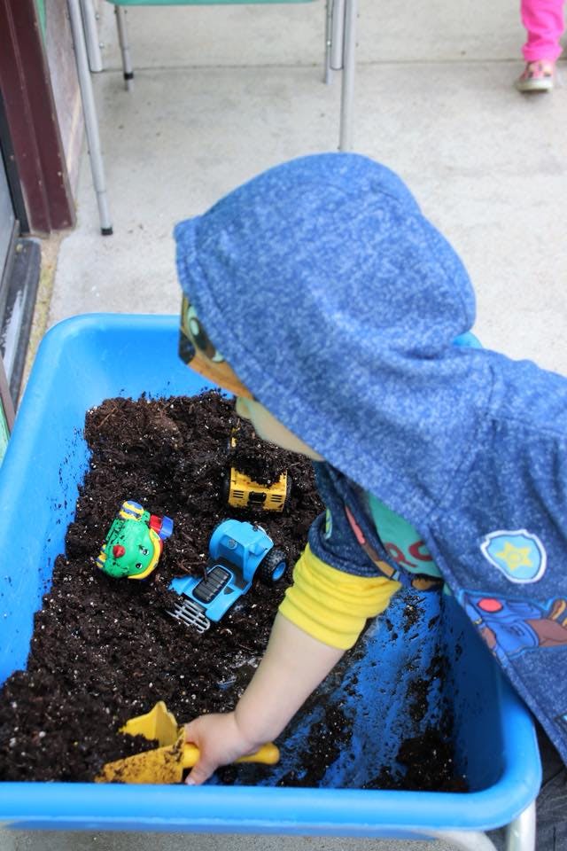 Child playing with toy trucks in a blue bin filled with dirt