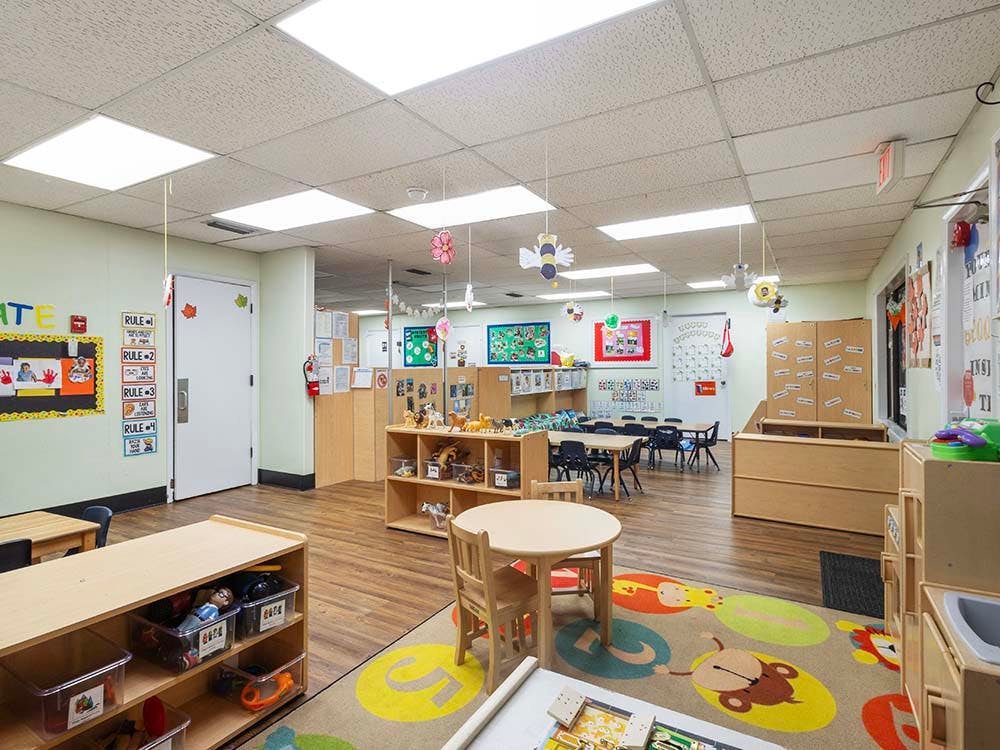 Empty classroom with small tables and chairs, shelves with toys, and wall decorations
