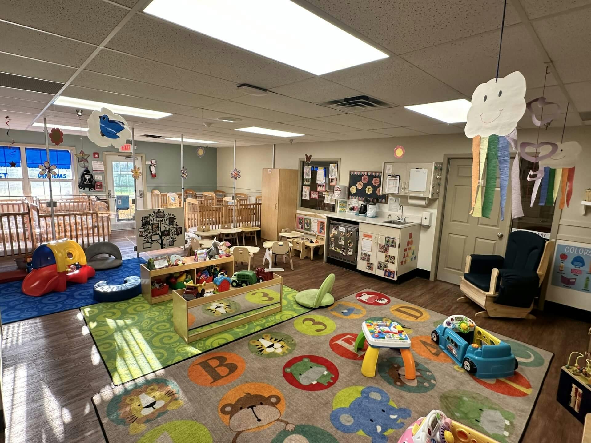 A colorful childcare classroom with cribs, play mats, and small furniture