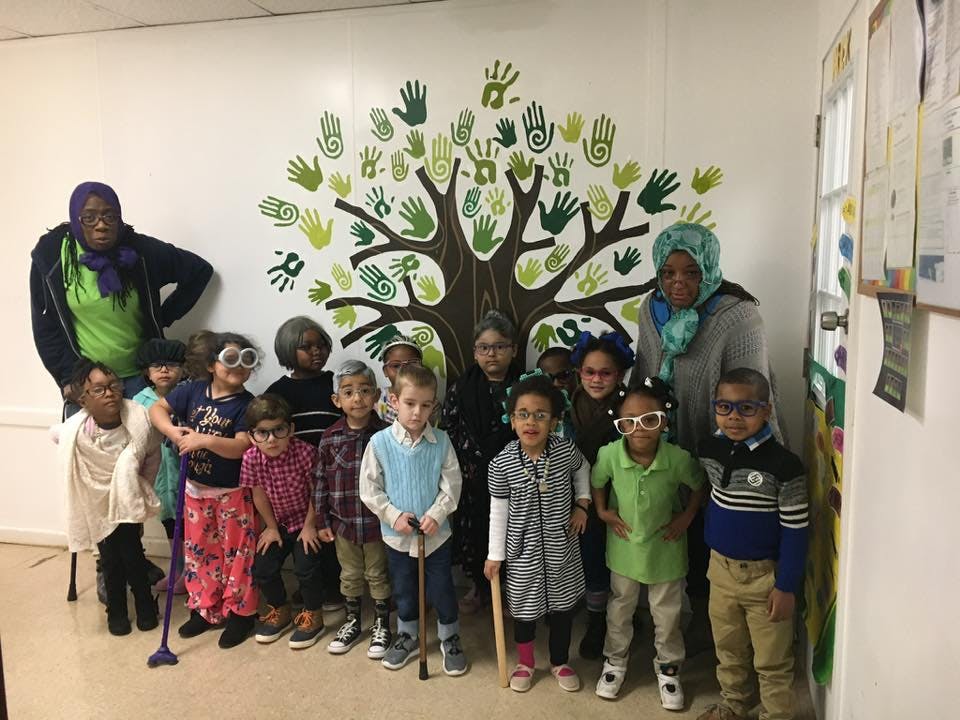 Two adults and a group of children posing in front of a tree mural