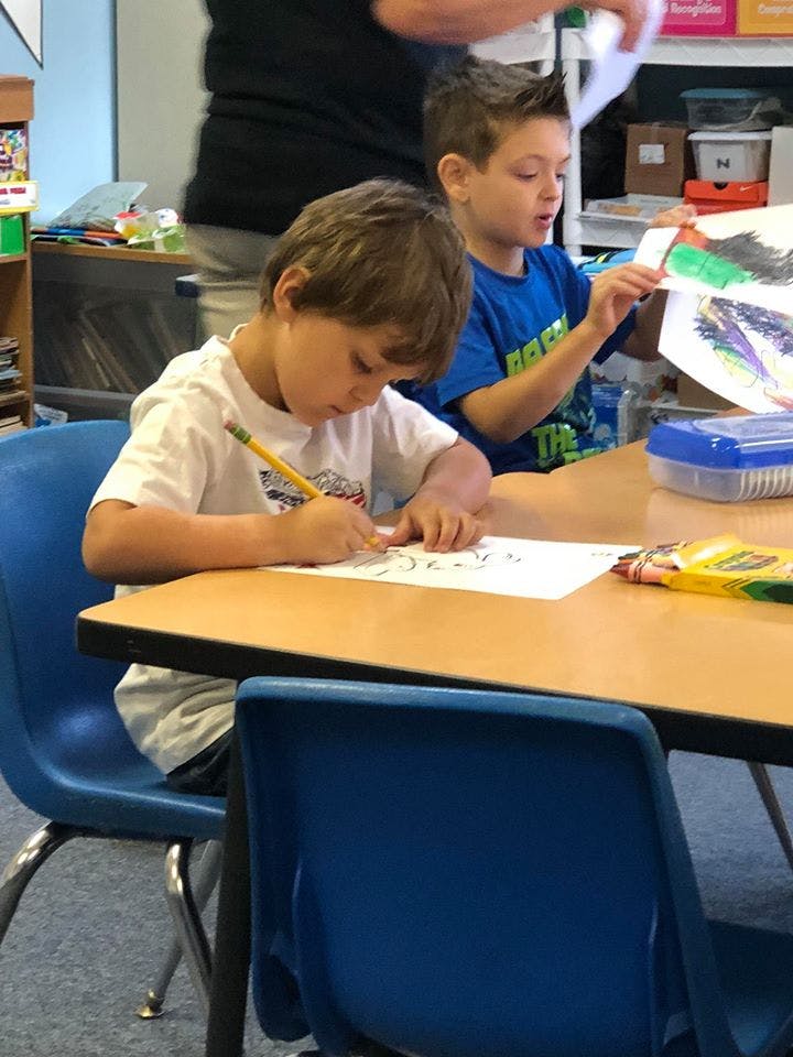 Child writing at a desk, another child holding up a drawing