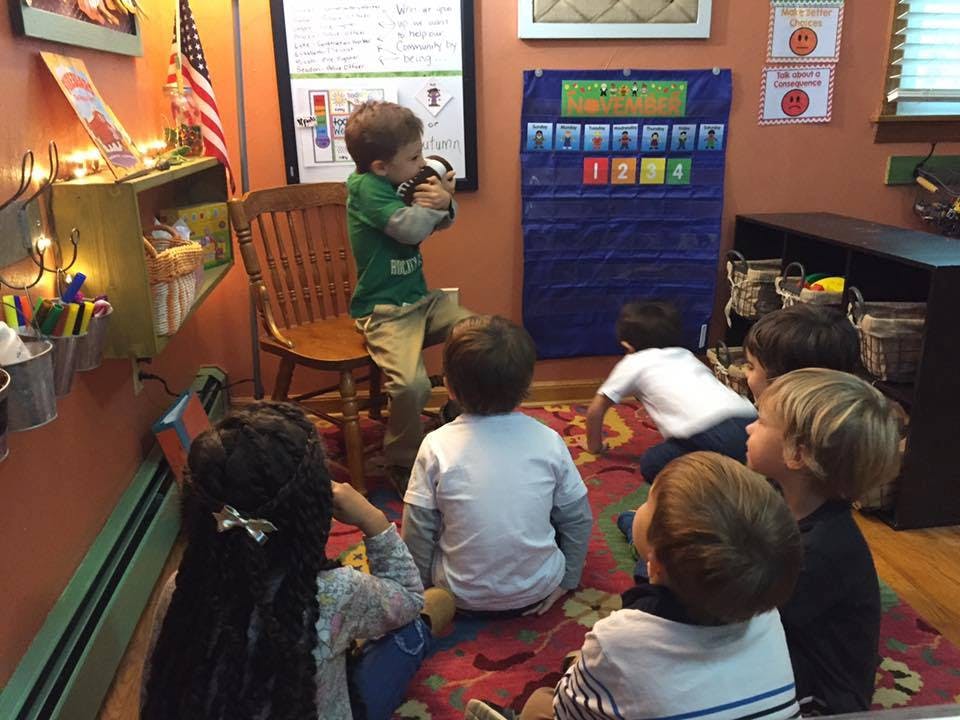 Child standing on a chair, speaking to other children sitting on a rug