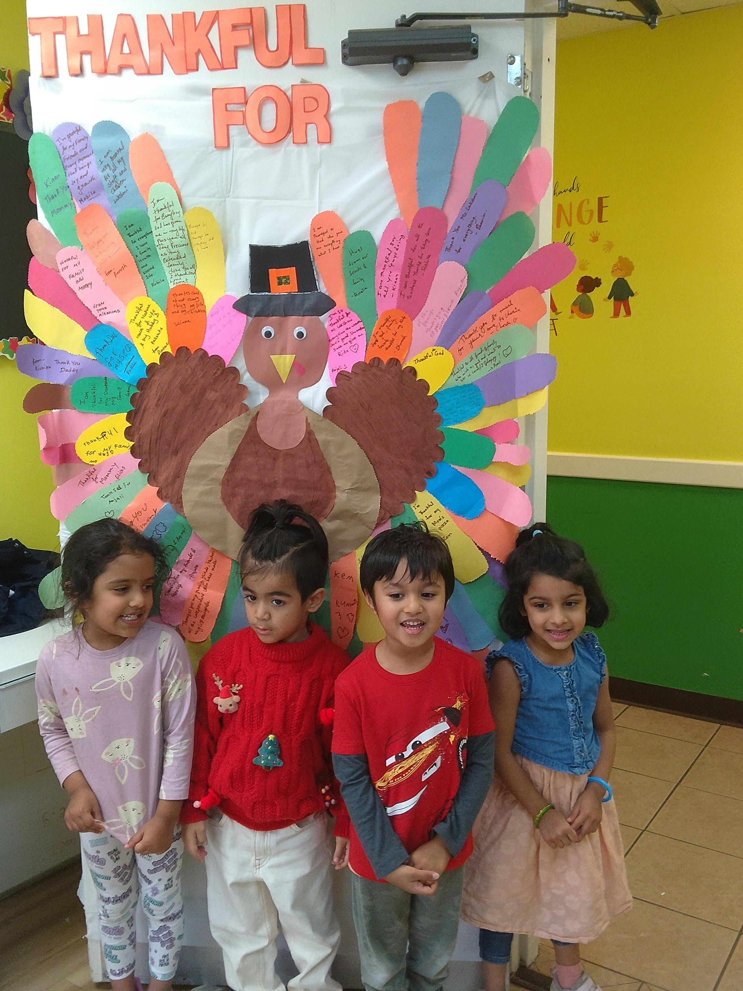 Four children standing in front of a paper turkey decoration with "THANKFUL FOR" above it