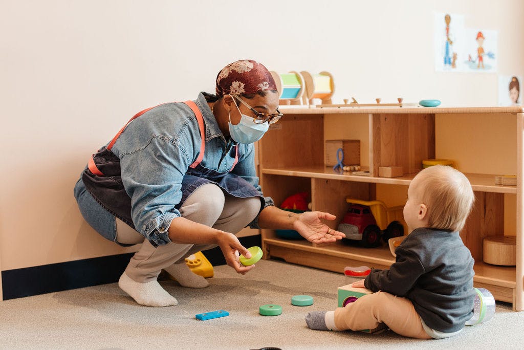 Person in mask crouching and offering a toy to a child sitting on the floor