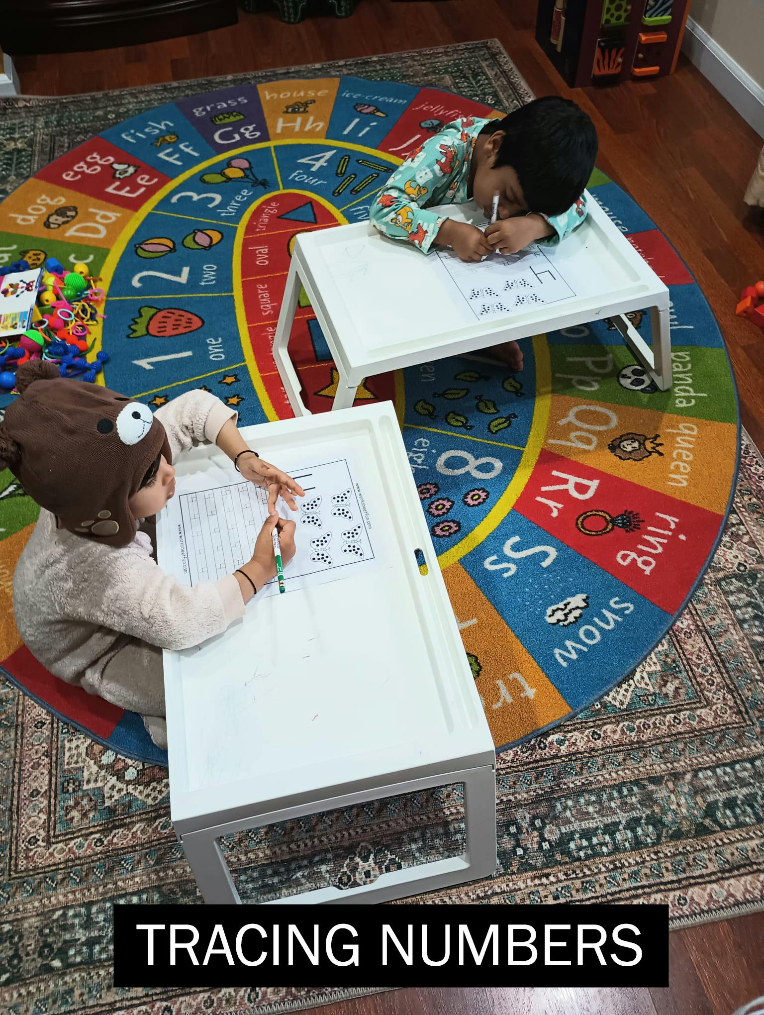 Two children on an alphabet rug, tracing numbers and letters at small tables