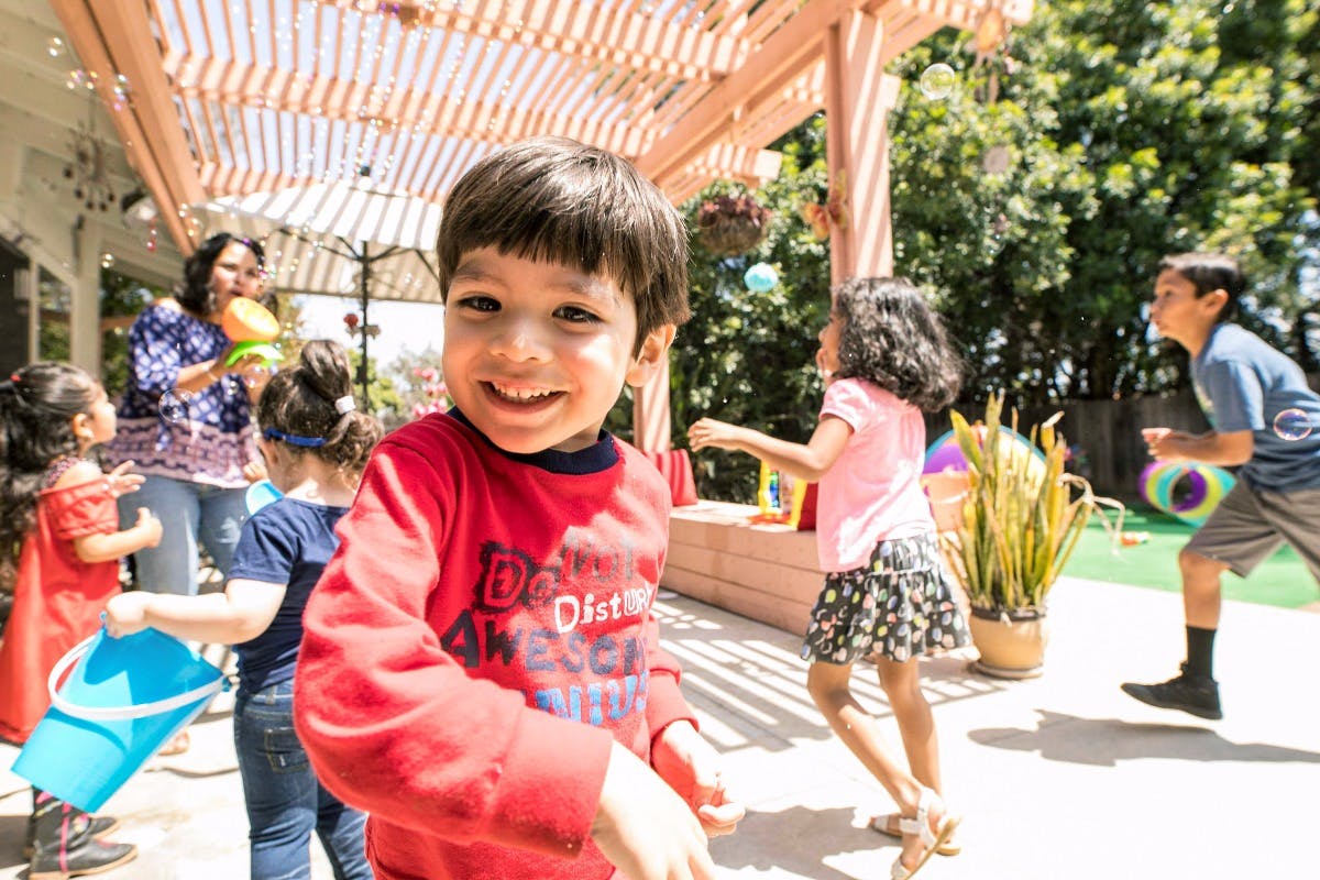 Child in red shirt smiling at camera with other children and adult playing in background