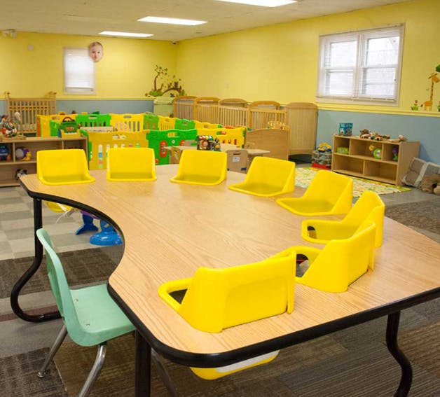 Classroom with a wooden table and yellow chairs, cribs in the background