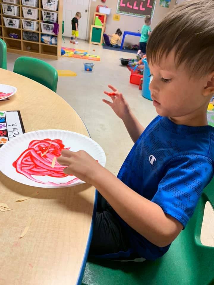 Child sitting at a table, working on a craft with red paint on a paper plate