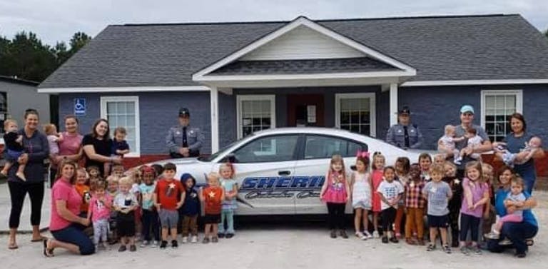 Group of adults and children posing with a sheriff's car in front of a building