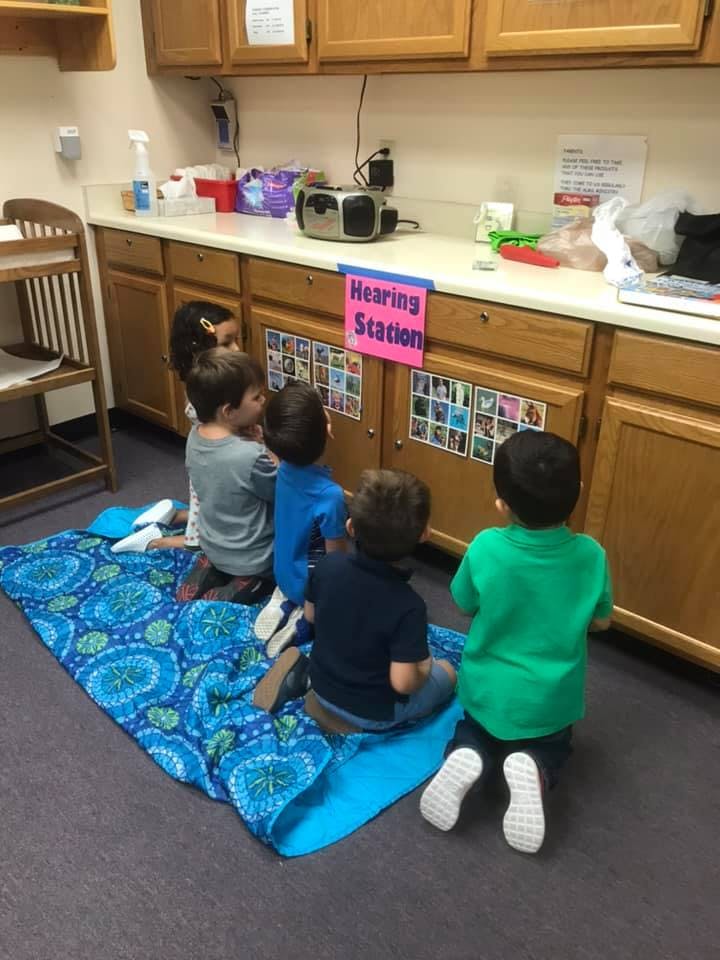 Children kneeling on a mat in front of a counter with a "Hearing Station" sign