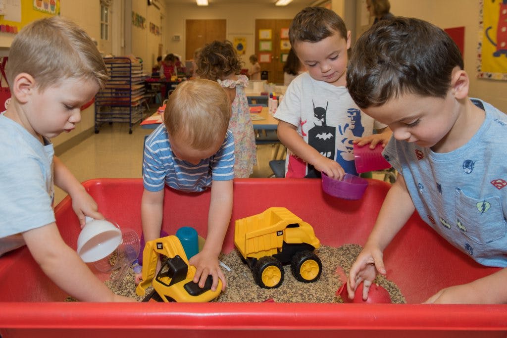 Four children playing with toy trucks and sand in a red bin