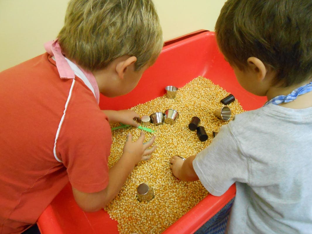 Two children playing with corn kernels and small objects in a red bin