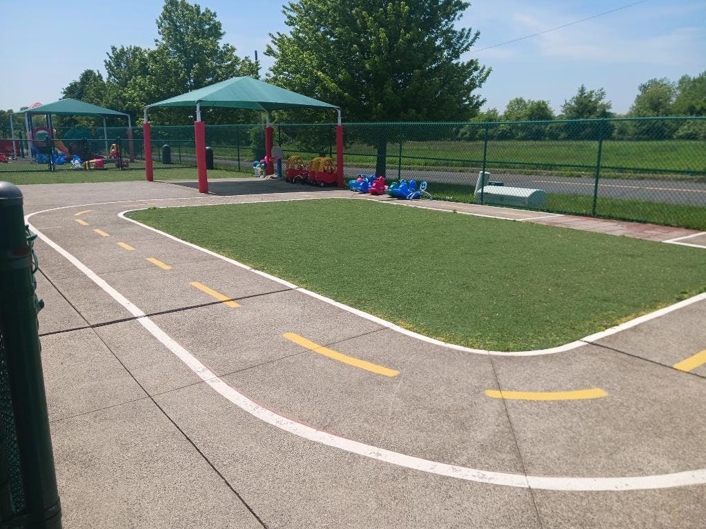 Outdoor play area with a paved track and green turf, ride-on toys under a canopy