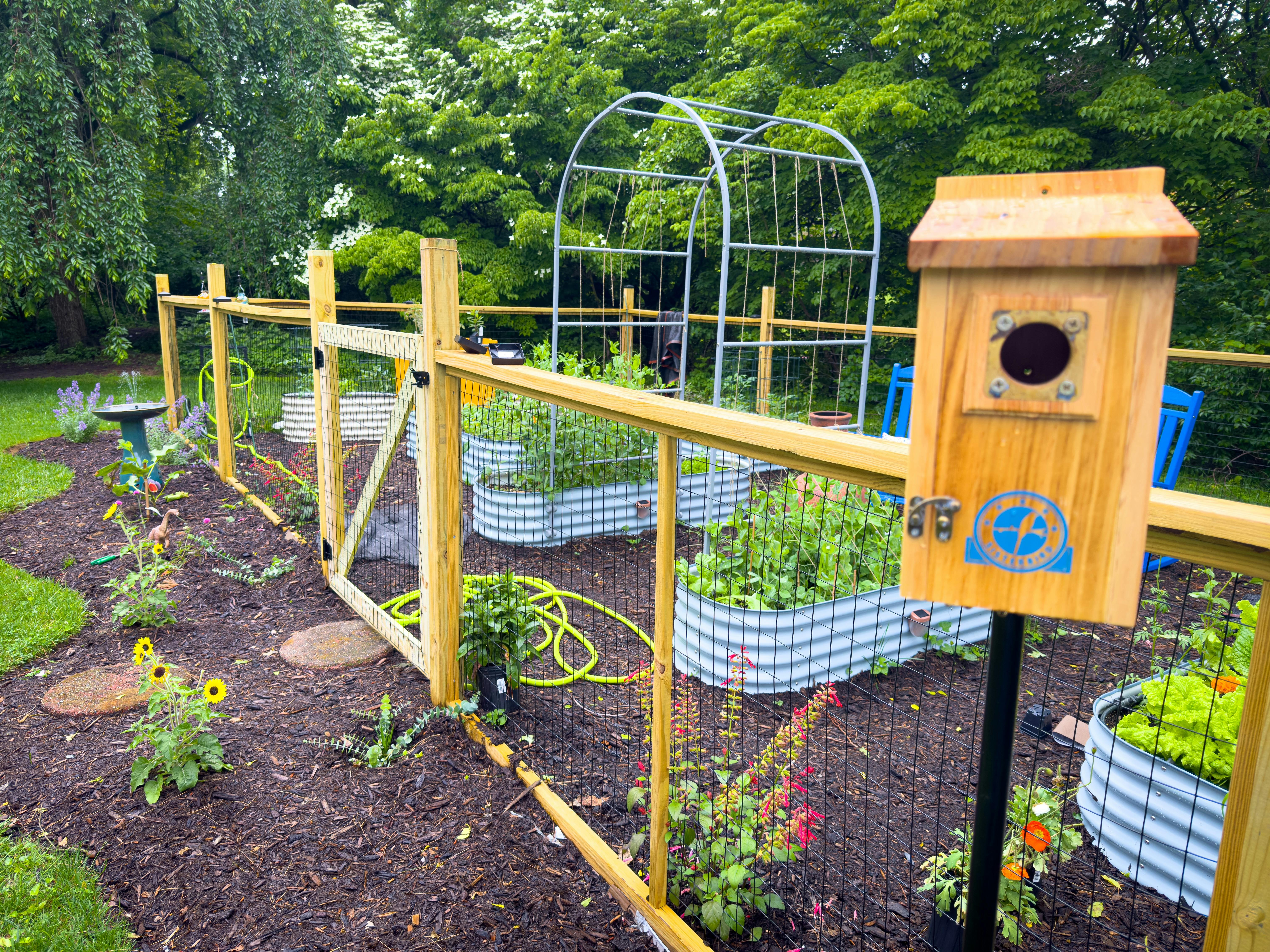 Wooden birdhouse on a fence post in front of a fenced vegetable garden