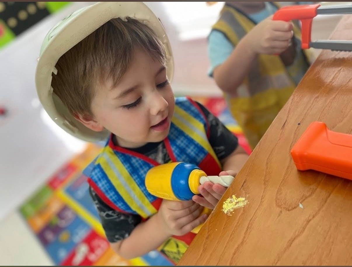 Child in a hard hat and vest holding a toy drill to a wooden surface