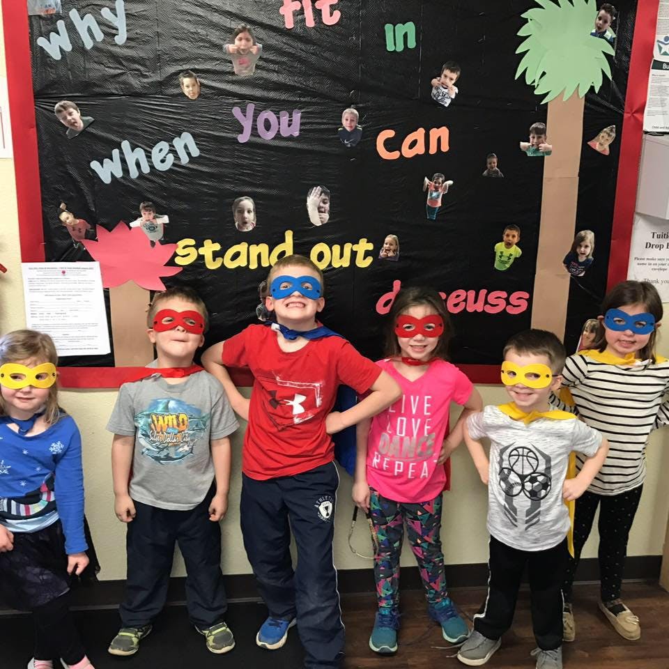 Five children wearing superhero masks and capes standing in front of a bulletin board