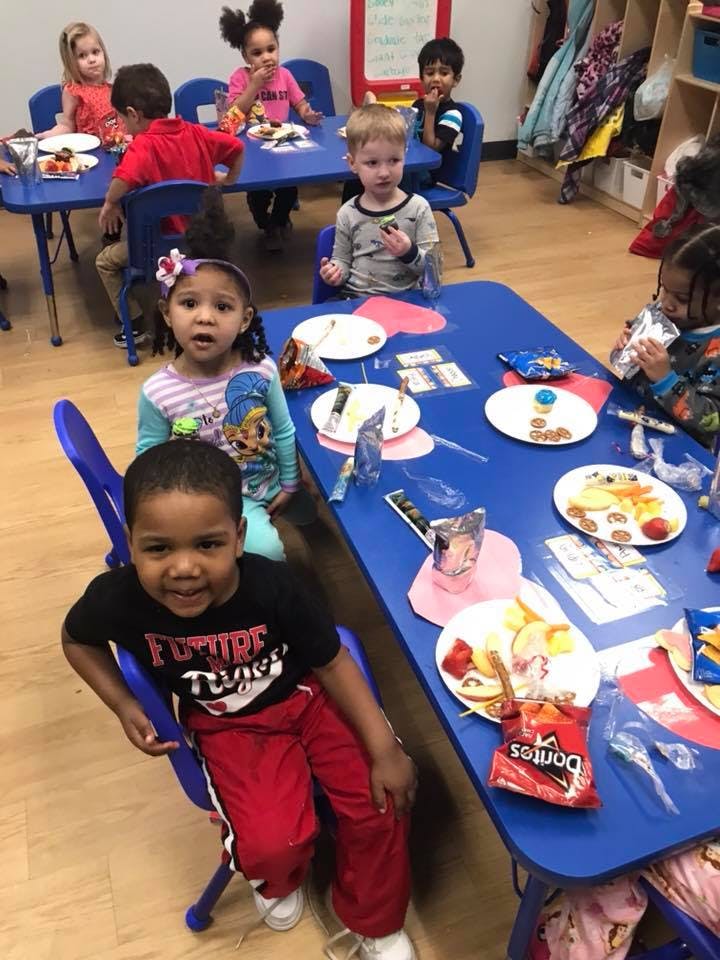 Children sitting at blue tables with plates of food in a classroom