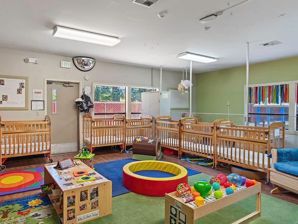 A brightly lit nursery room with several wooden cribs and colorful play mats