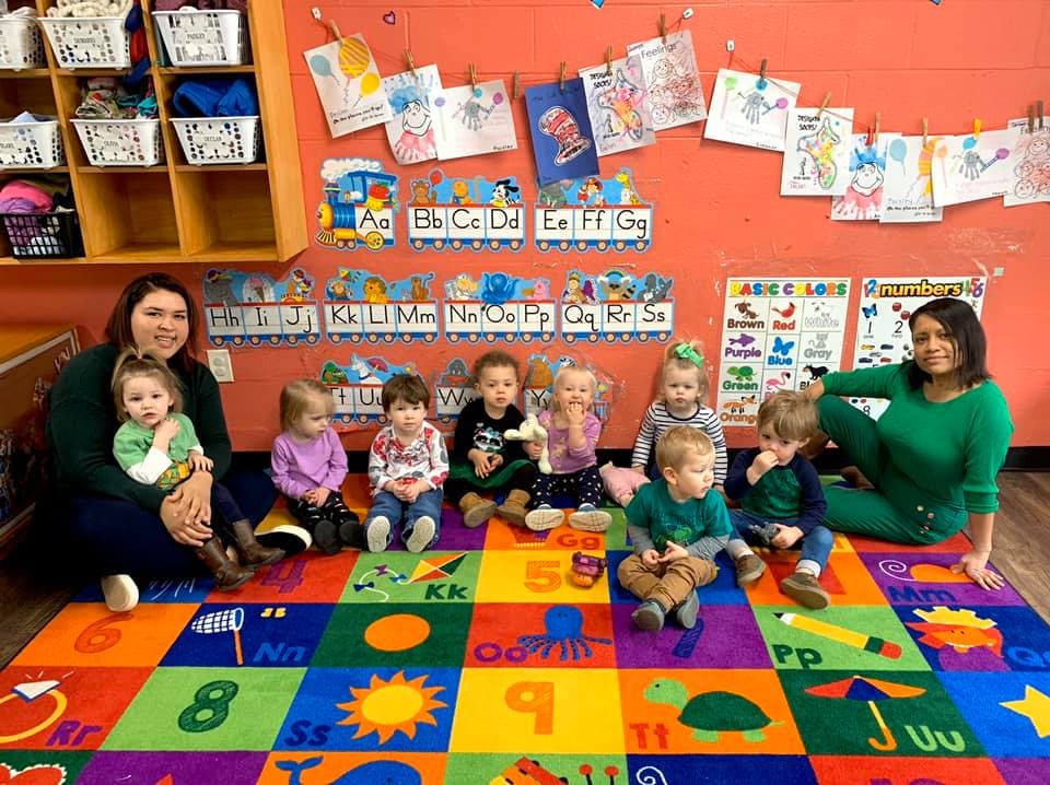 Two adults with several young children sitting on a colorful rug in a classroom