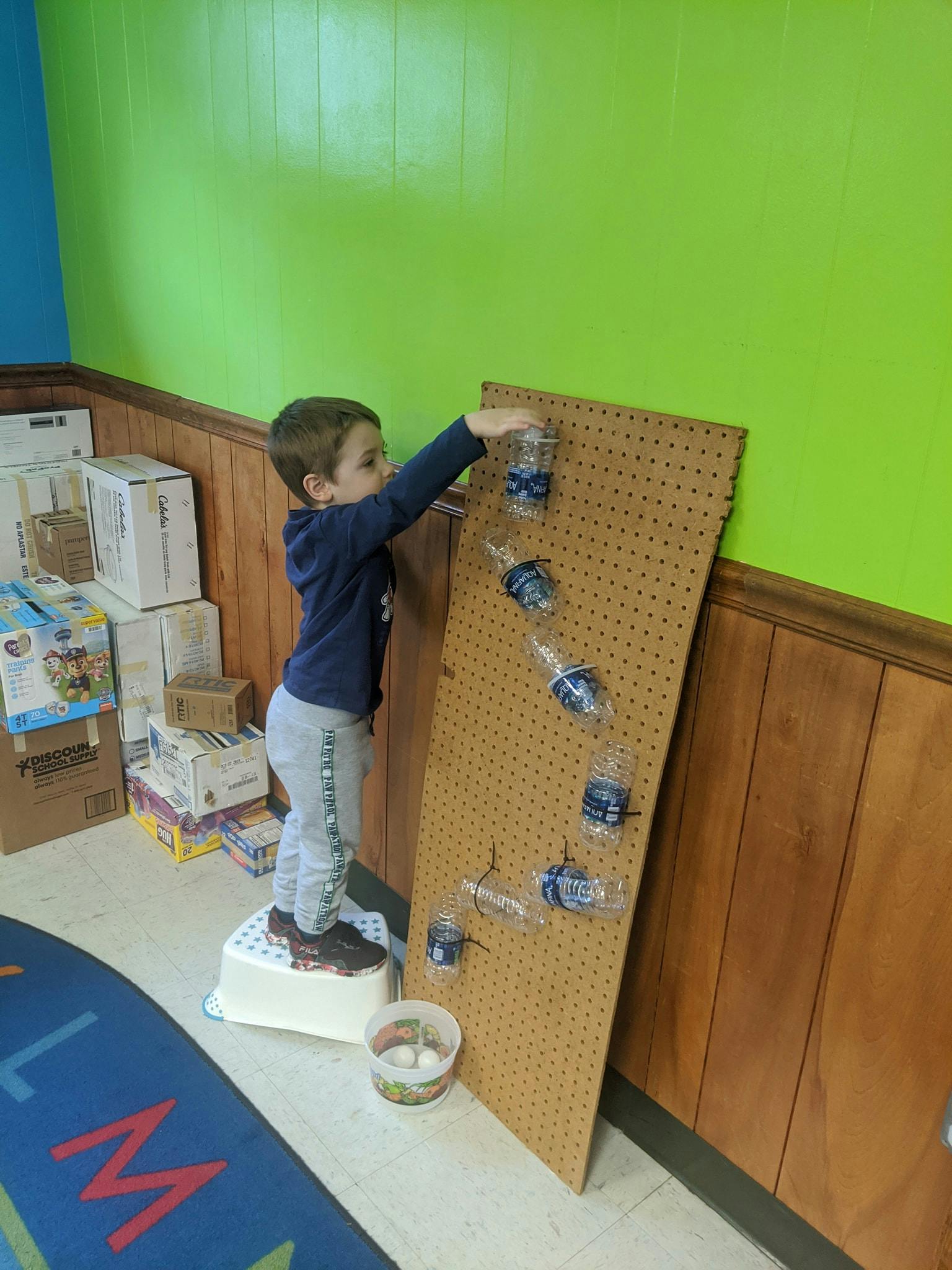 Child on a step stool reaching for a plastic bottle on a pegboard