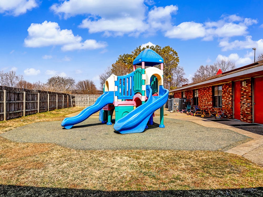 A colorful playground structure with slides on a padded surface next to a brick building
