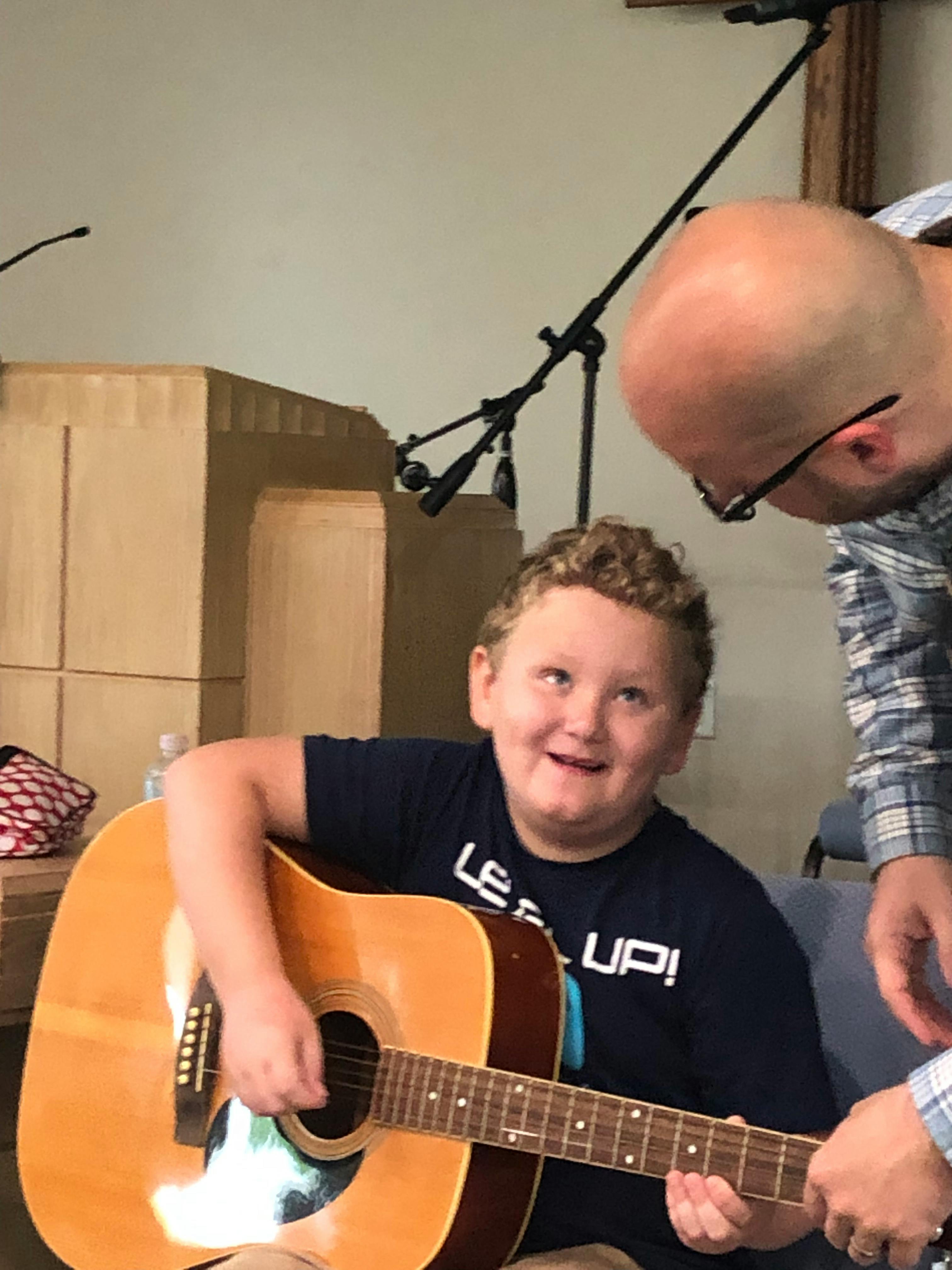 Child playing an acoustic guitar with an adult nearby
