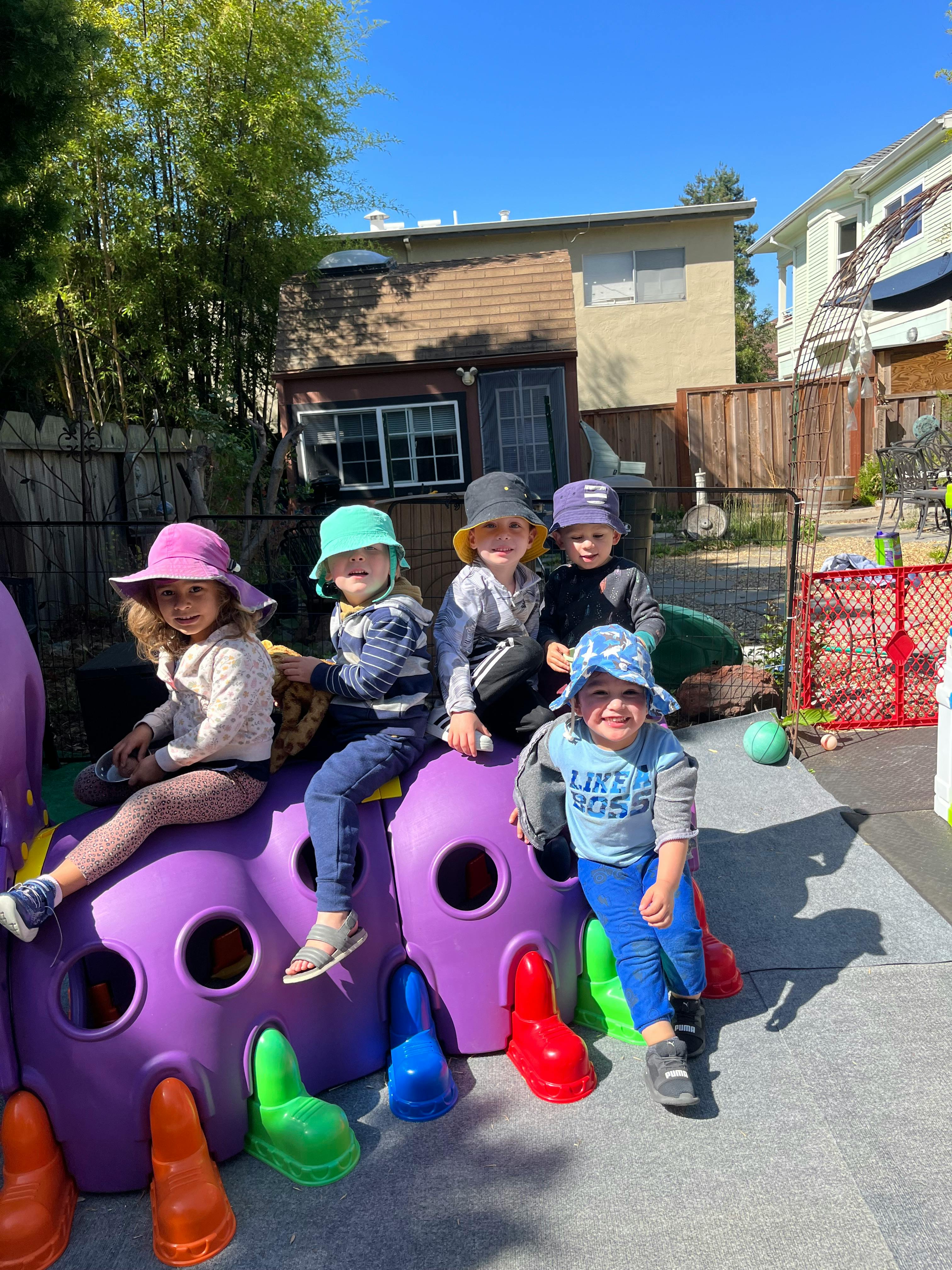 Five children in hats sitting on a purple caterpillar playground toy