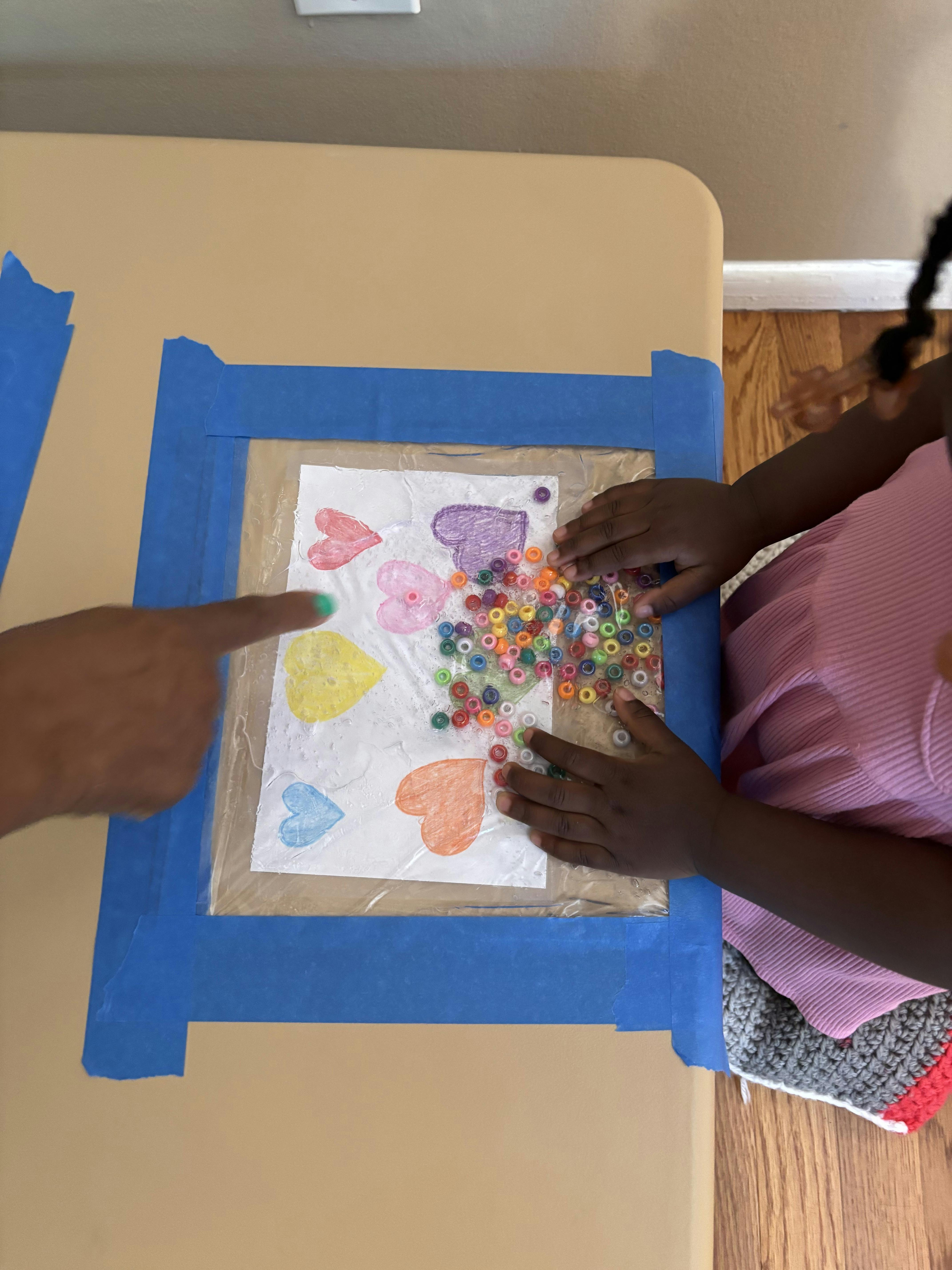 Child and adult playing with plastic beads on a taped paper with heart drawings
