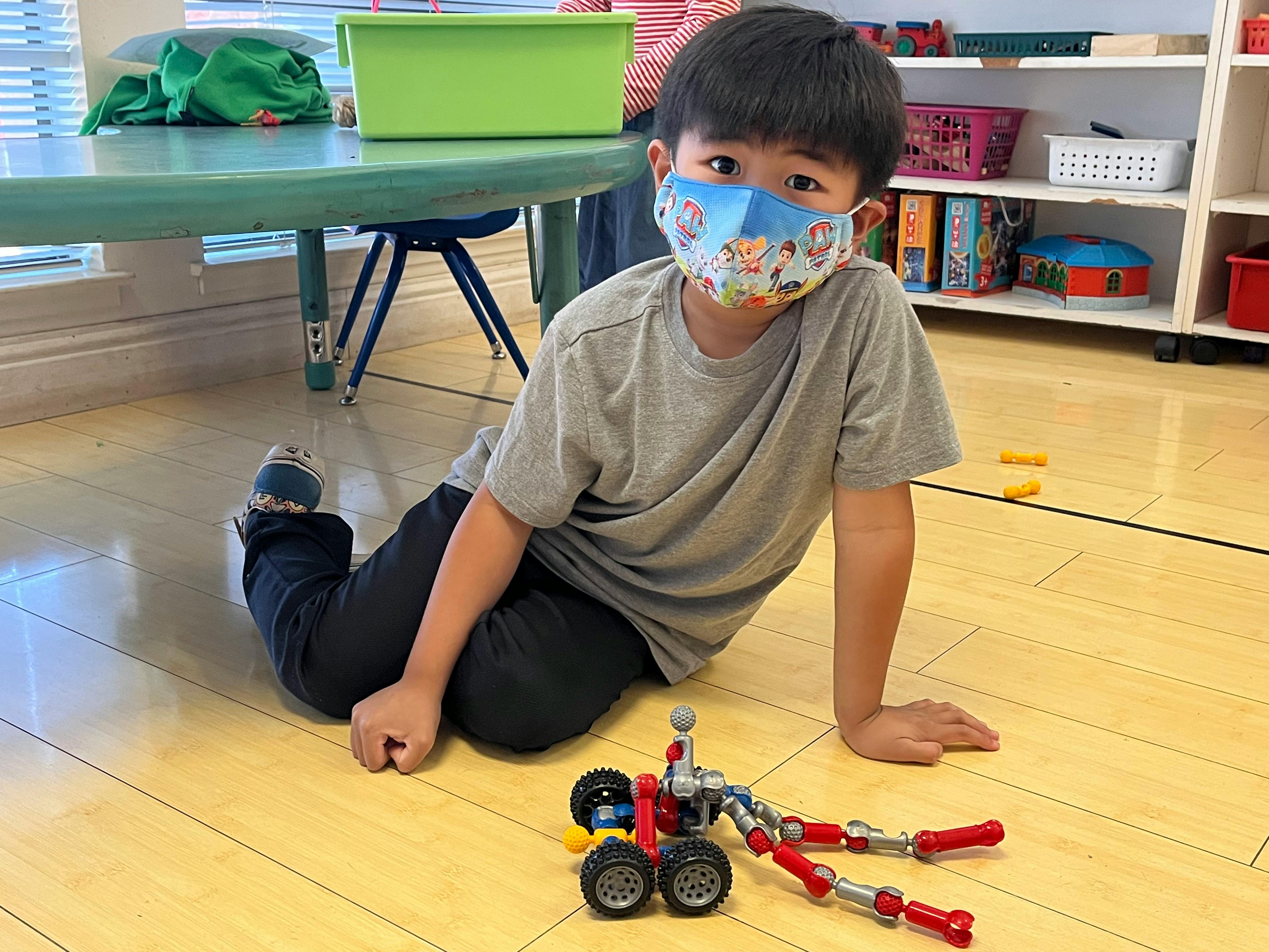 Child in a face mask sitting on the floor with a toy