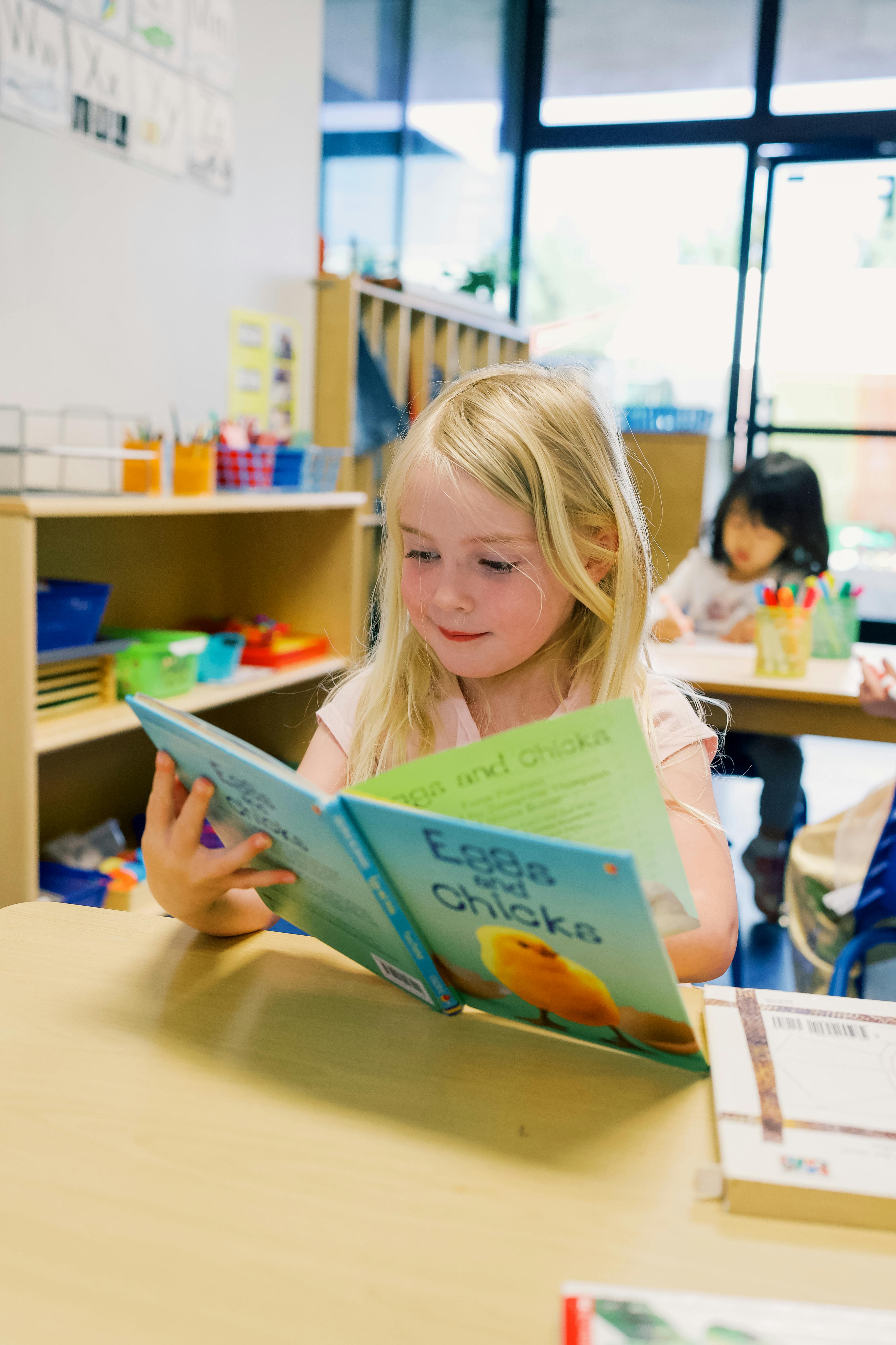 Child with blonde hair reading a book at a desk in a classroom