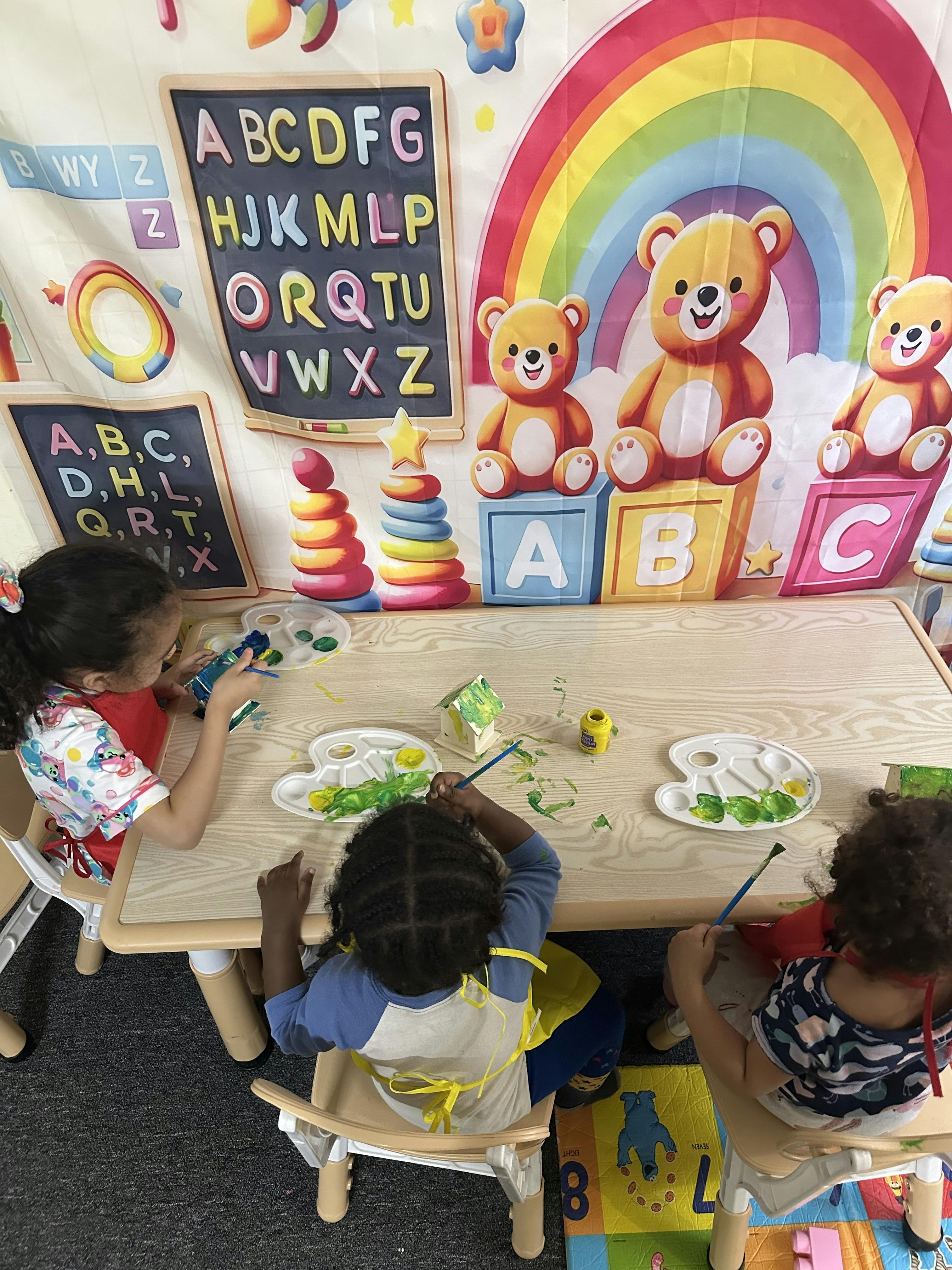 Three children painting at a table in a classroom with alphabet decorations