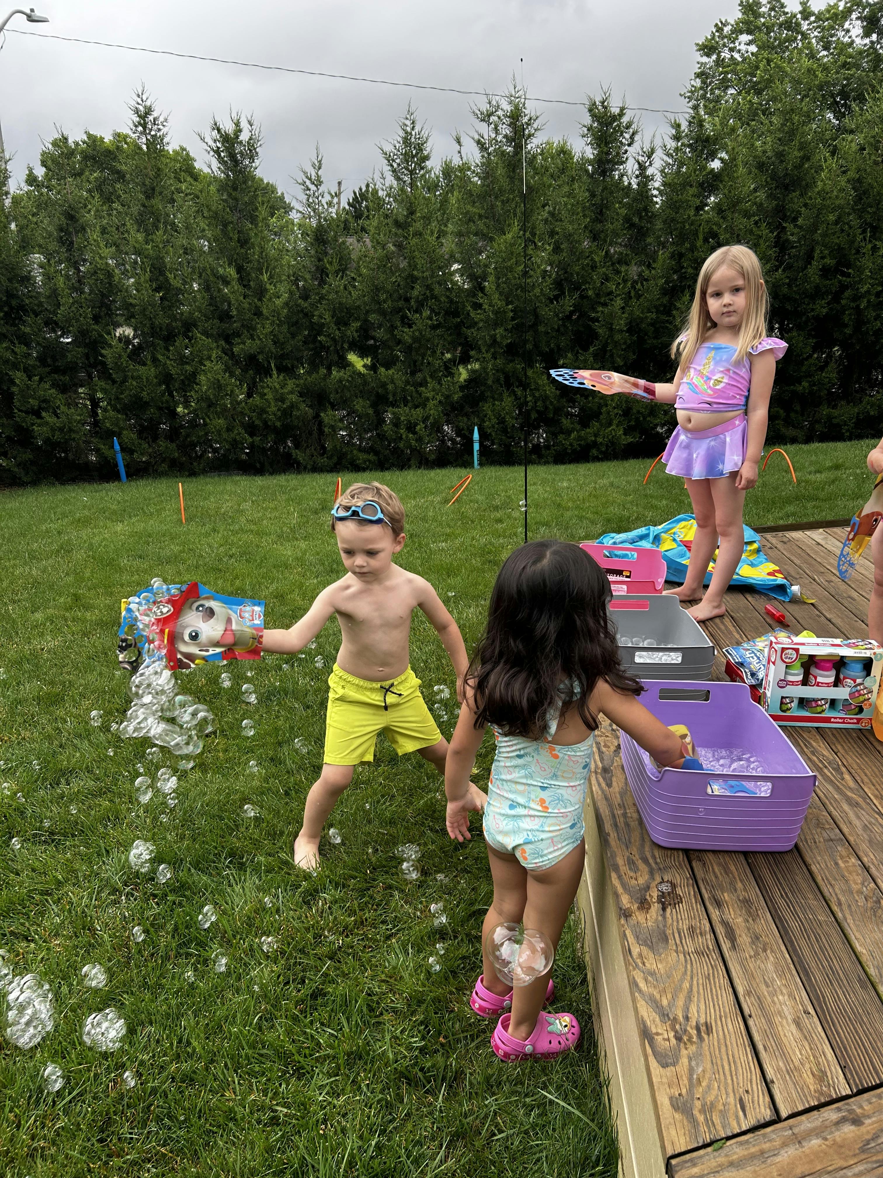 Three children playing with bubbles in a grassy yard next to a wooden deck