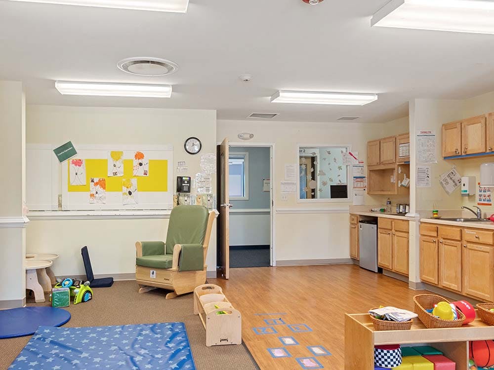 Classroom with a rocking chair, child art on a bulletin board, and a small kitchen area