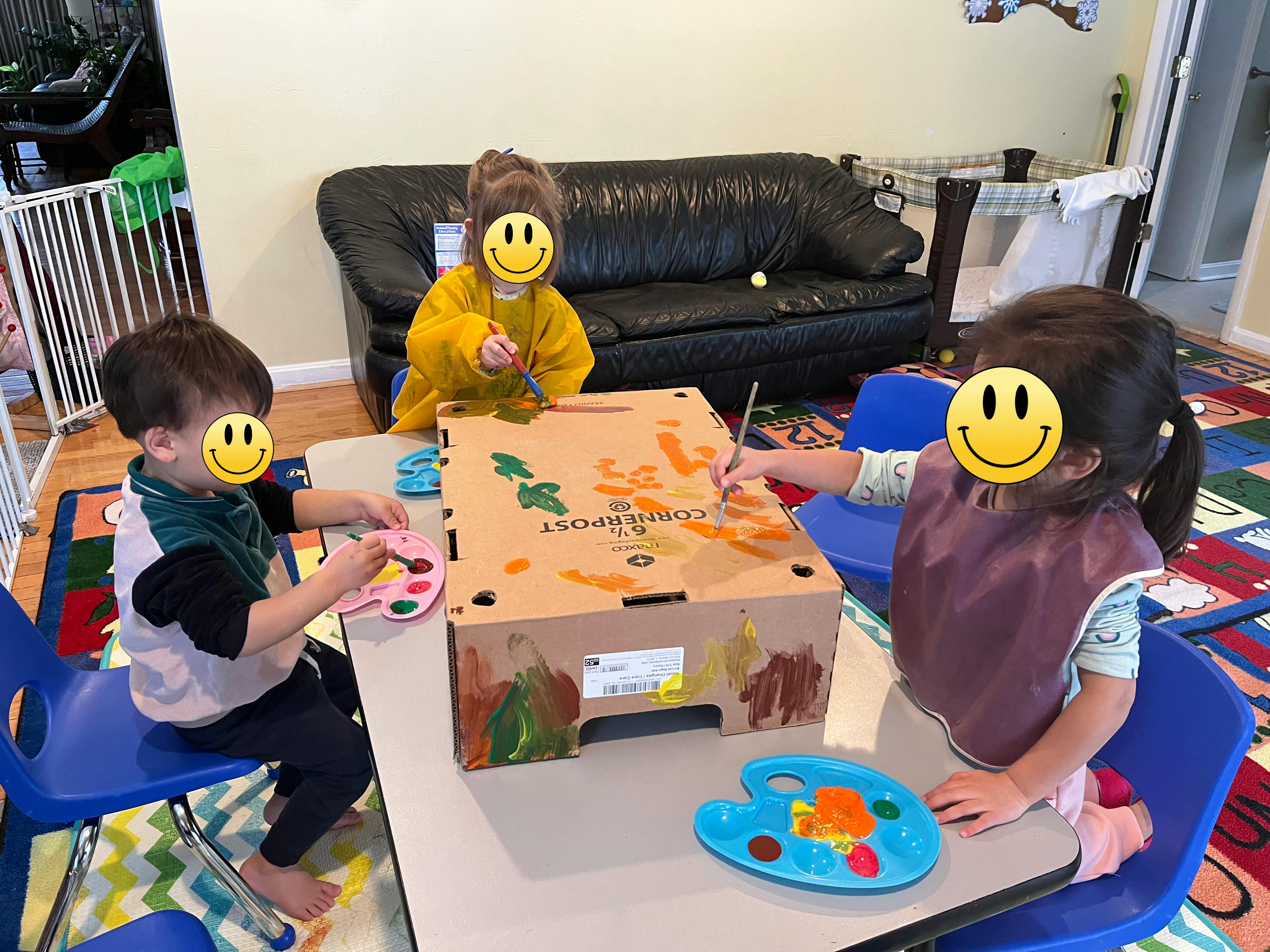 Three children wearing aprons painting on a cardboard box at a table