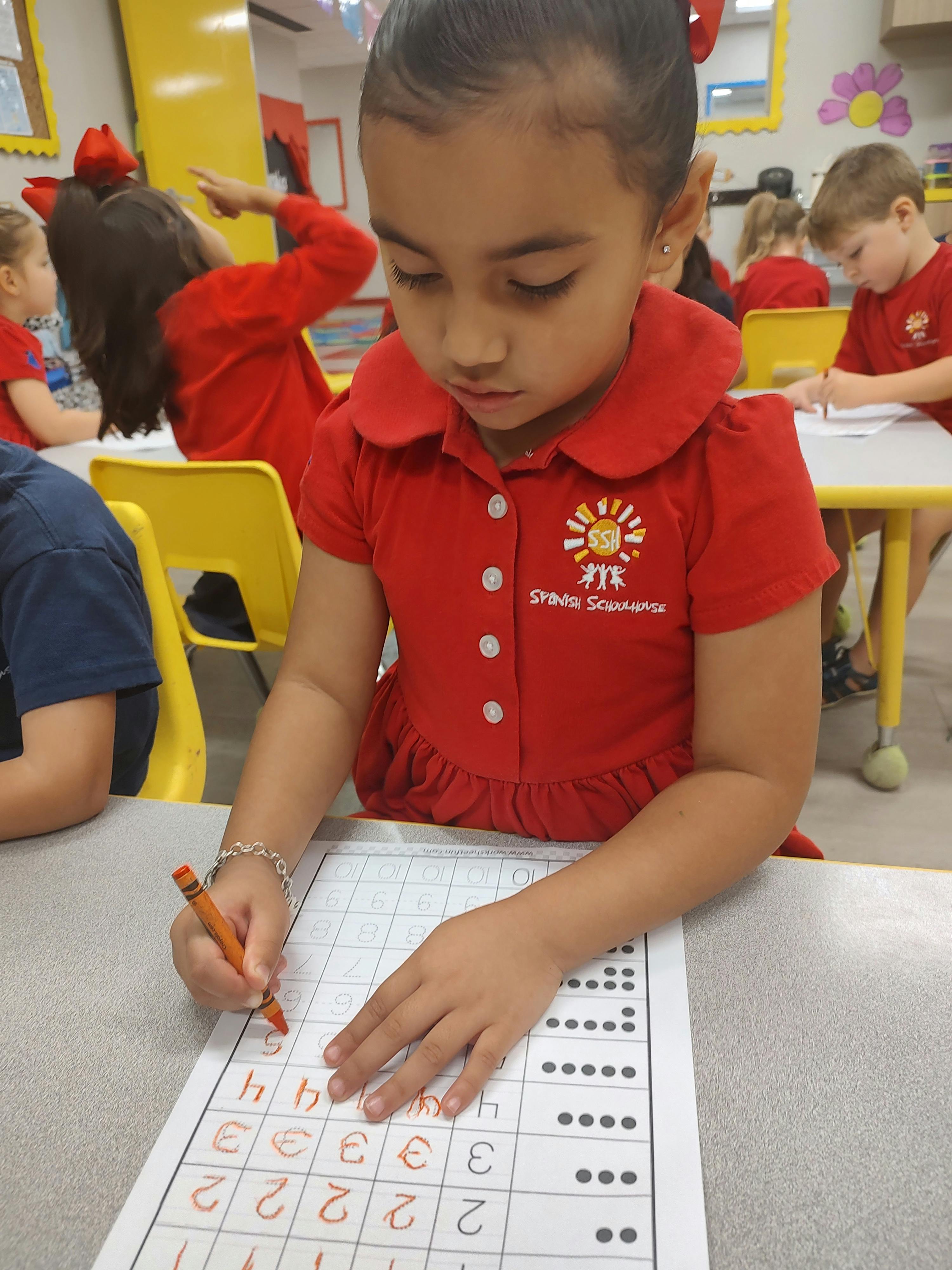 Child in red shirt writing on a worksheet in a classroom