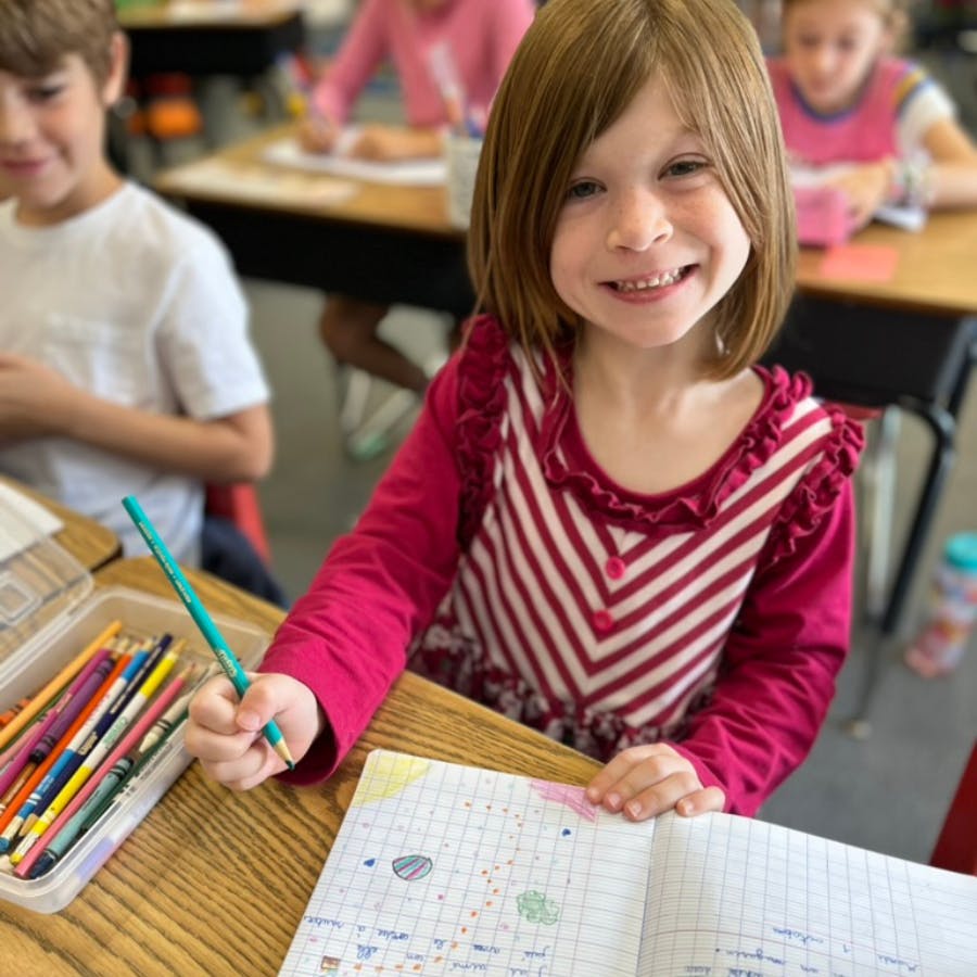 Child holding a pencil and writing in a notebook at a desk in a classroom