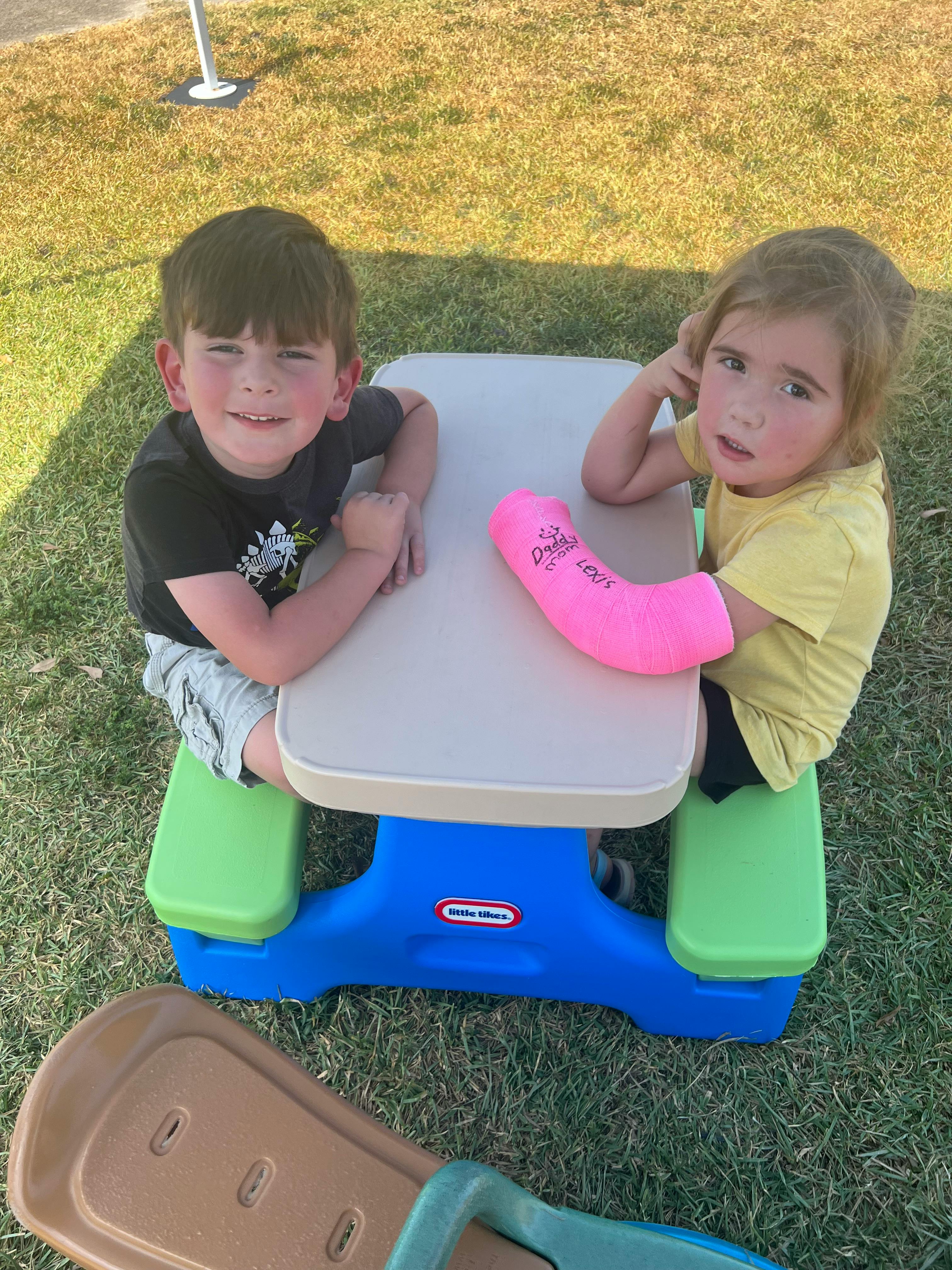 Two children sitting at a small picnic table, one with a pink cast
