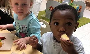 Two children at a table, one eating a piece of fruit