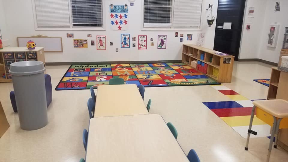 Empty classroom with small tables and chairs, colorful rug, and shelves
